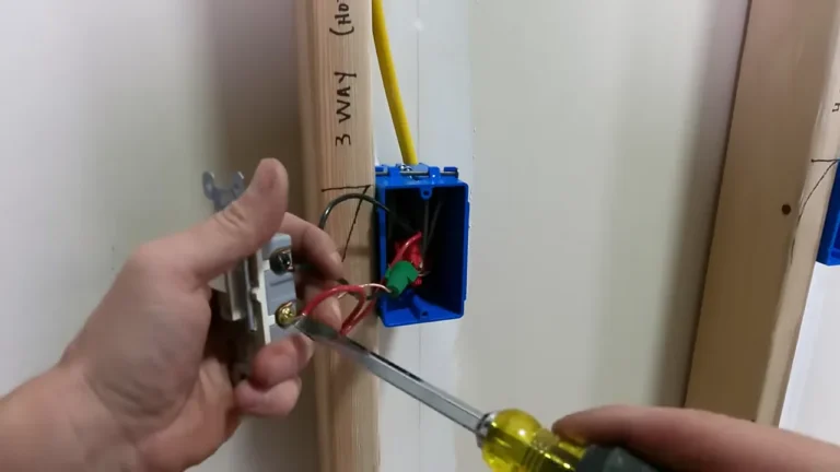 Hands wiring a 3-way switch, tightening a red traveller wire onto the switch's brass screw beside an open blue wall box