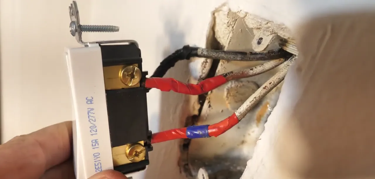 Replacement 3-way switch held at an open wall box with the old wires marked in black, red and blue electrical tape so each wire lands back on the right screw